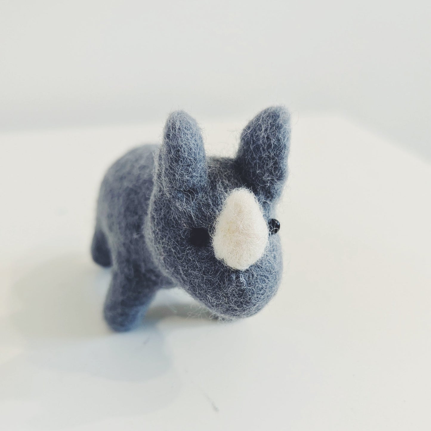 A small felted wool toy rhino, resembling a French bulldog, in a dark blue-gray color with a white snout, standing on a plain white surface.