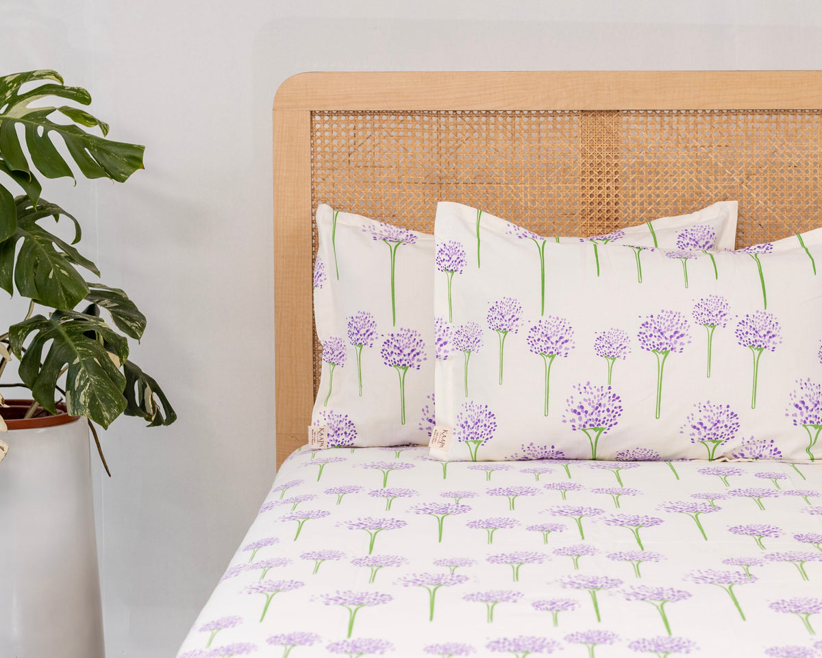 A neatly made bed with a yellow floral quilt, two matching pillow shams, a white quilted mattress topper, and a beige striped throw blanket, set in a warm, sunlit bedroom with a wooden headboard.