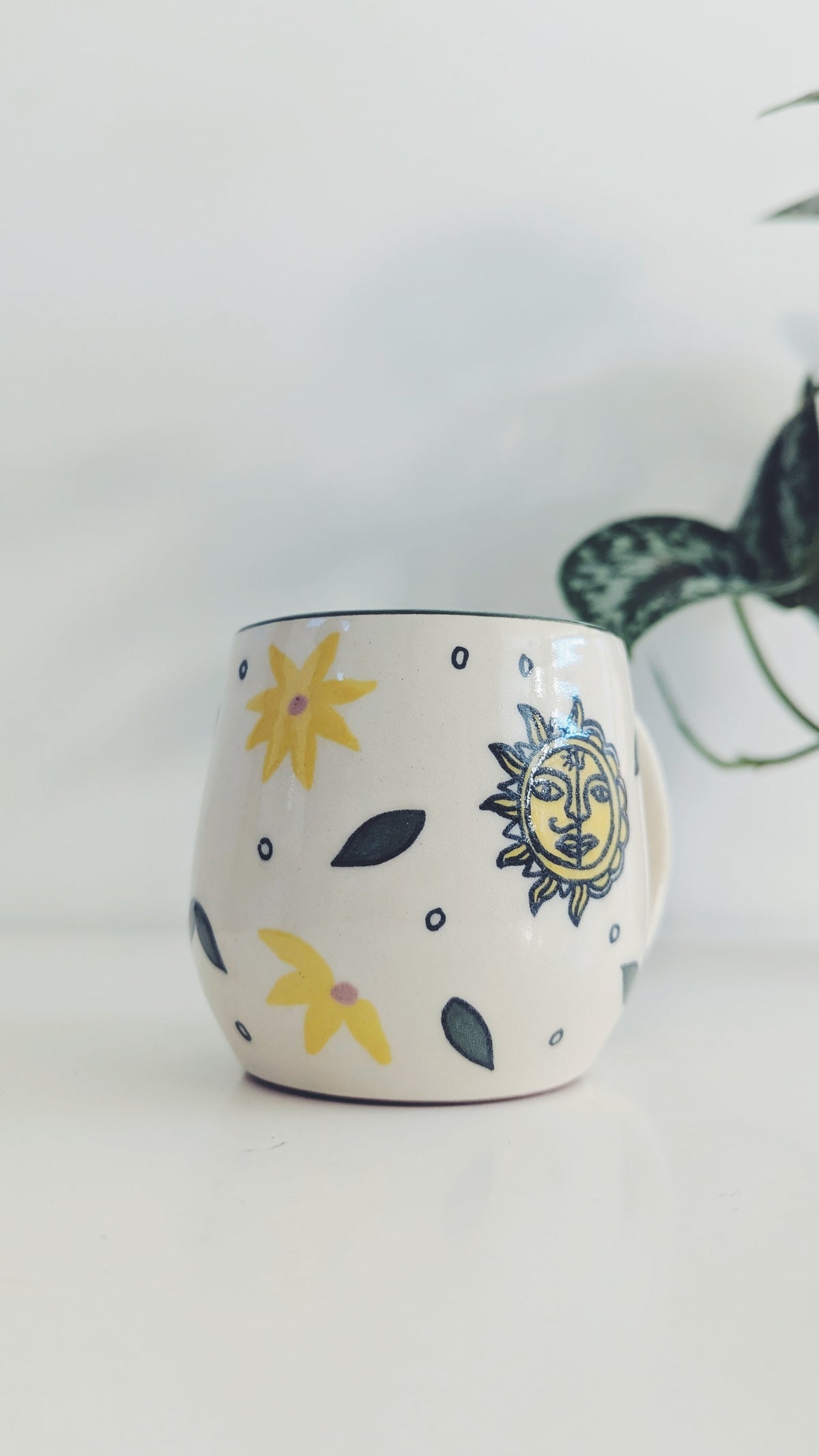 A white ceramic mug decorated with hand-painted in mithila art yellow flowers, a smiling sun face, and dark gray leaves, set against a bright white background with a blurred green plant in the background.