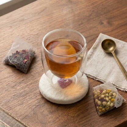 A clear double-walled glass cup filled with amber tea sits on a round white coaster on a wooden table. Two small tea bags are placed nearby, one on a folded cream cloth with a gold spoon, creating a cozy tabletop tea scene.