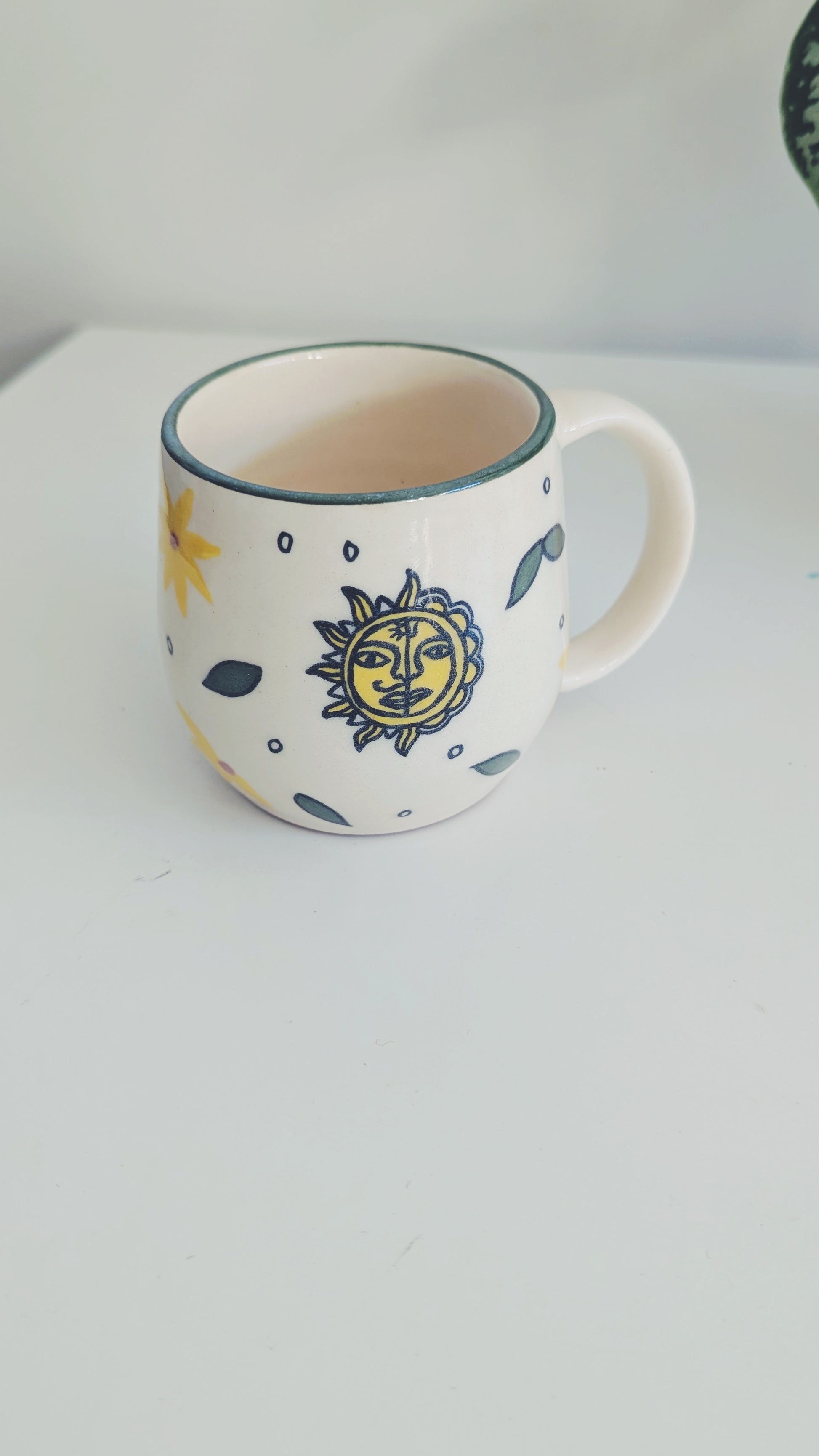 A white ceramic mug with a yellow sun face mithila art design and blue accents, sitting on a white surface against a white background.