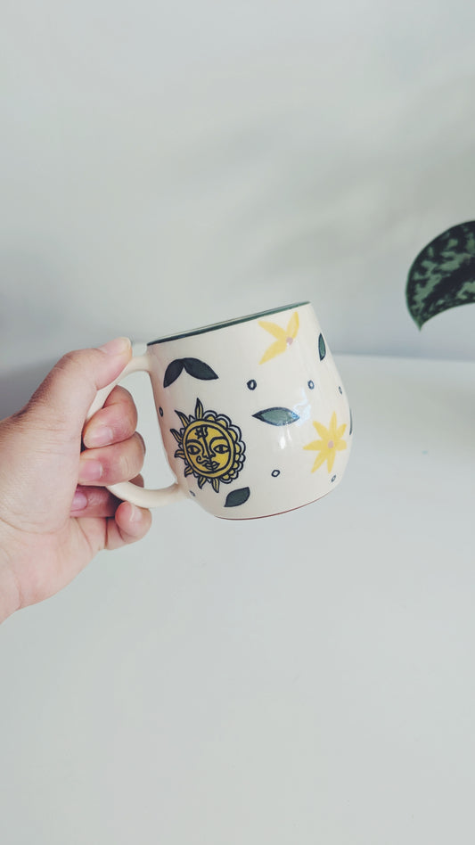 A hand holding a cream-colored mithila art ceramic mug decorated with a yellow sun, yellow flowers, and dark gray leaves, set against a minimalist light gray background.