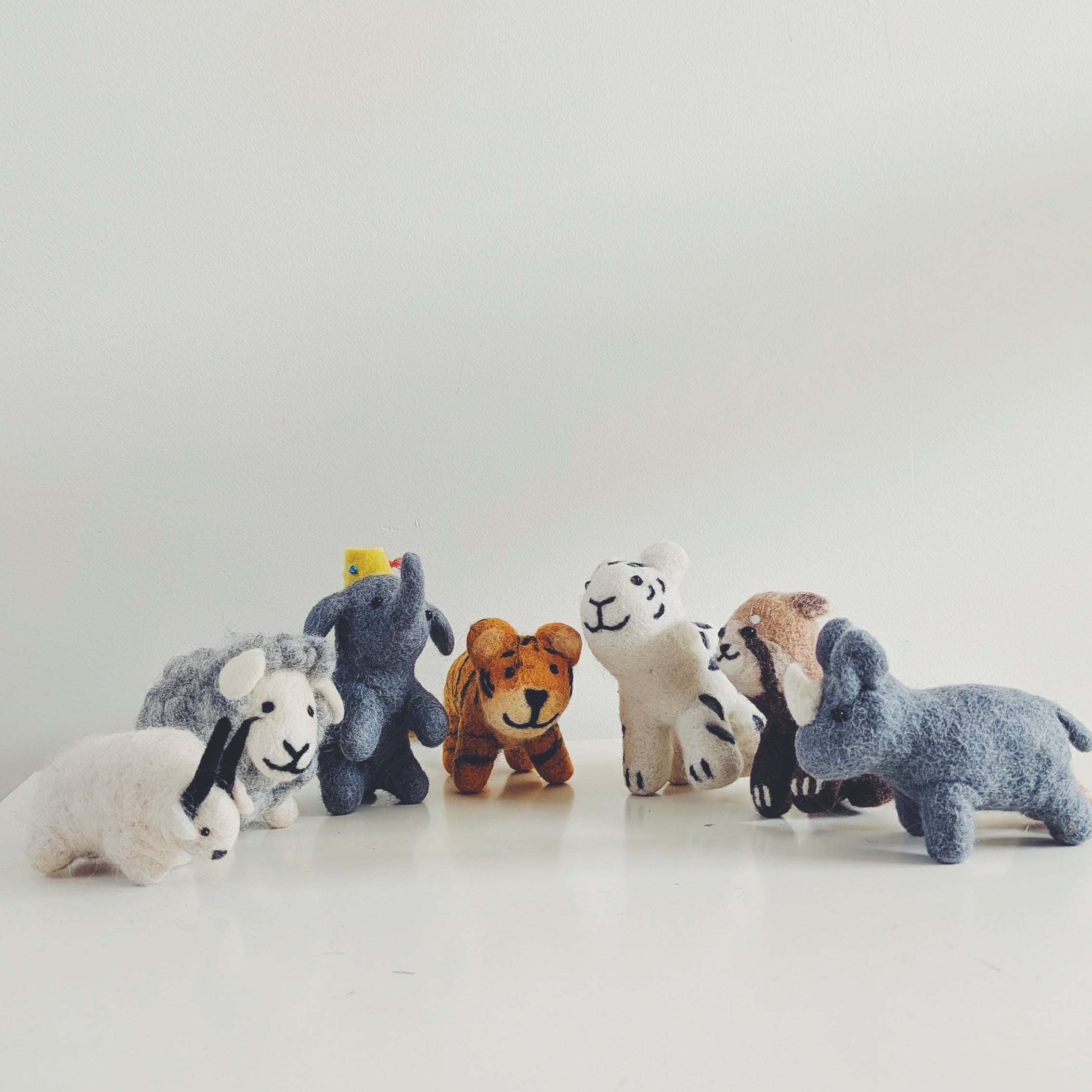 A row of small animal felt figurines, including a tiger, red panda, himalayan goat, elephant, yak, rhino, sheep, and white tiger, arranged on a white surface against a plain white background in Pascoe vale australia