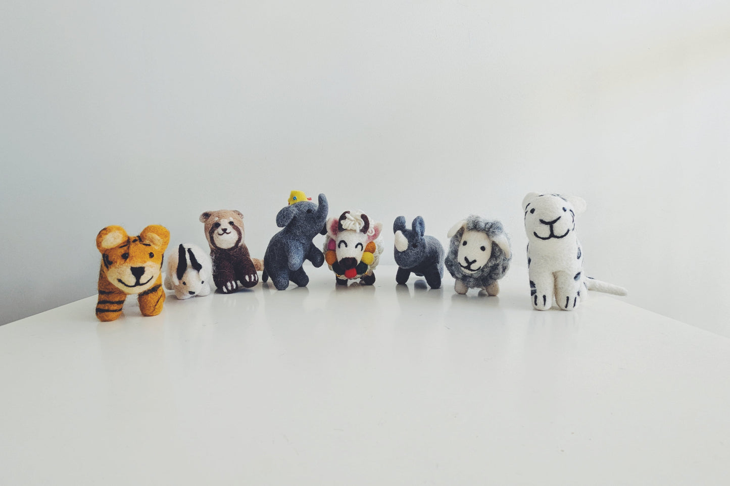 A row of small animal felt figurines, including a tiger, red panda, himalayan goat, elephant, yak, rhino, sheep, and white tiger, arranged on a white surface against a plain white background in Pascoe vale australia