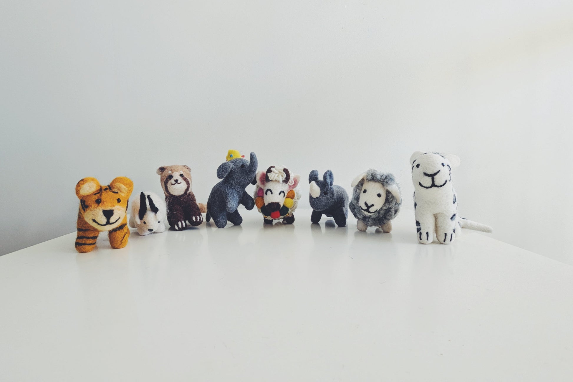 A row of small animal felt figurines, including a tiger, red panda, himalayan goat, elephant, yak, rhino, sheep, and white tiger, arranged on a white surface against a plain white background in Pascoe vale australia