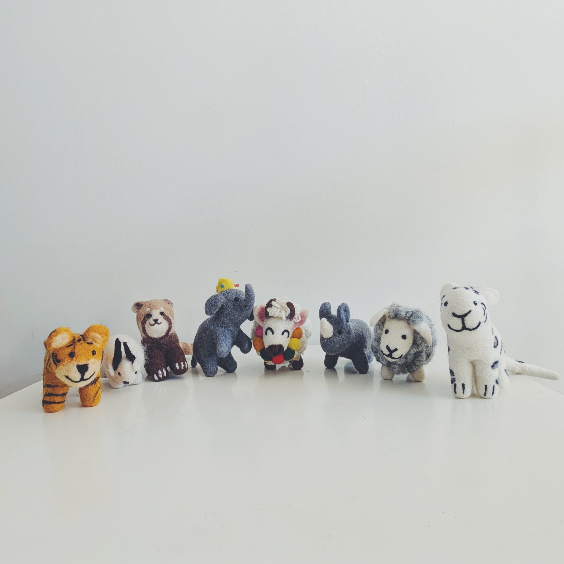 A row of small animal felt figurines, including a tiger, red panda, himalayan goat, elephant, yak, rhino, sheep, and white tiger, arranged on a white surface against a plain white background in Pascoe vale australia