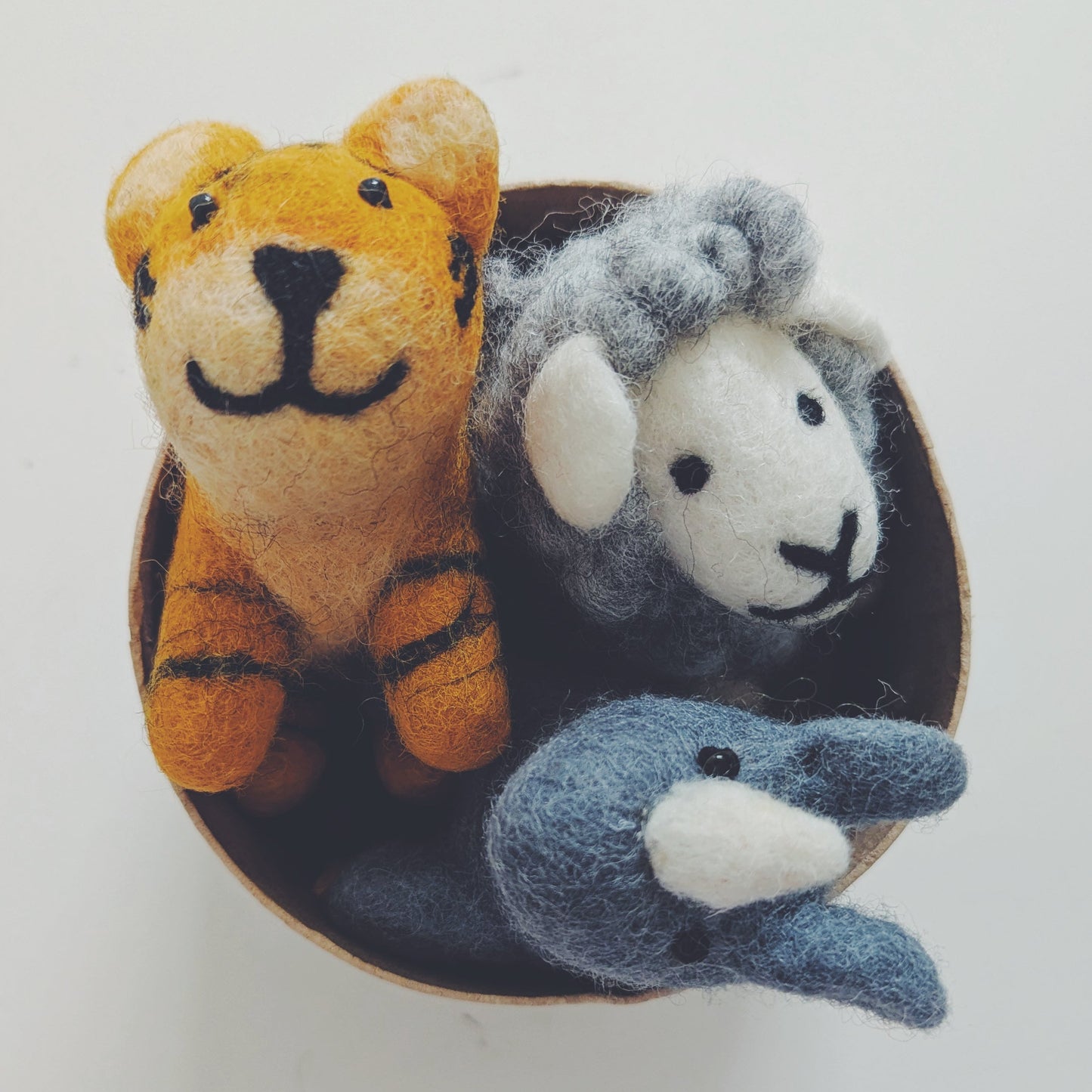 Three small felt animal toys arranged inside a round dark dish on a plain light background: an orange tiger, a gray sheep, and a blue-gray rhino