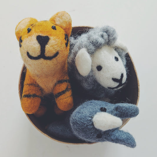 Three small felt animal toys arranged inside a round dark dish on a plain light background: an orange tiger, a gray sheep, and a blue-gray rhino