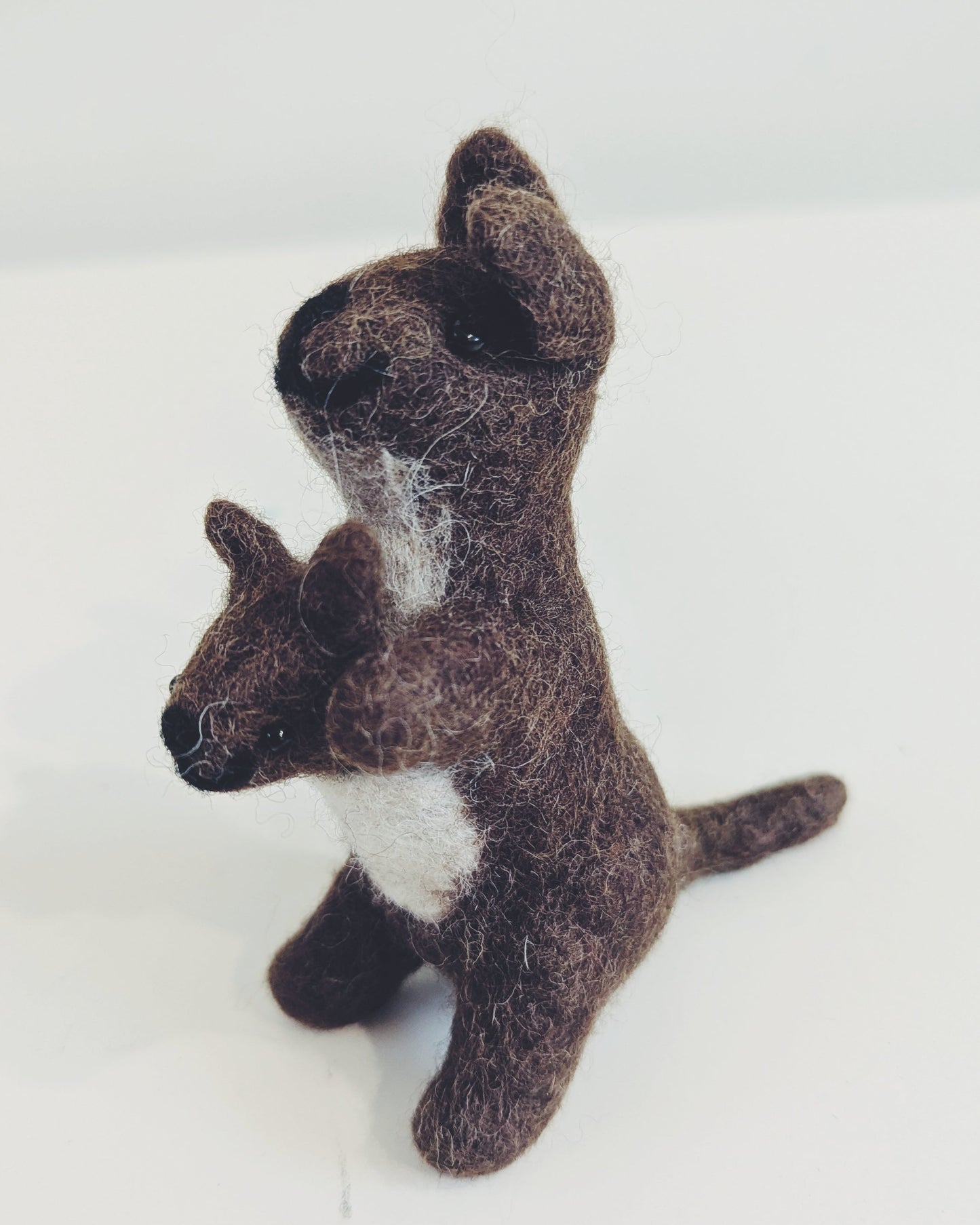 A small felted wool toy kangaroo holding a matching joey, crafted in dark brown with a white chest patch, standing on a plain white surface.