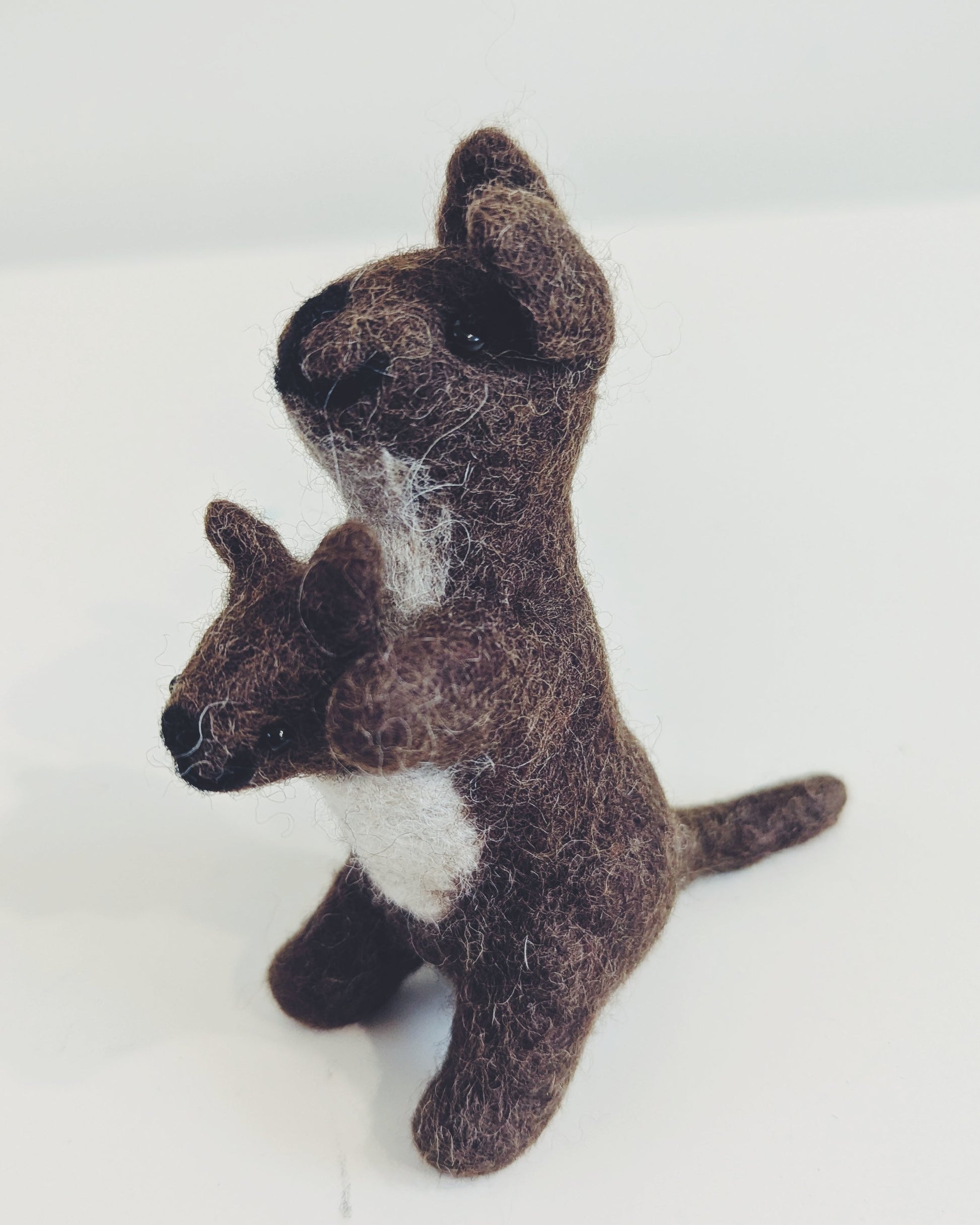 A small felted wool toy kangaroo holding a matching joey, crafted in dark brown with a white chest patch, standing on a plain white surface.