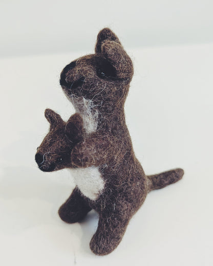 A small felted wool toy kangaroo holding a matching joey, crafted in dark brown with a white chest patch, standing on a plain white surface.