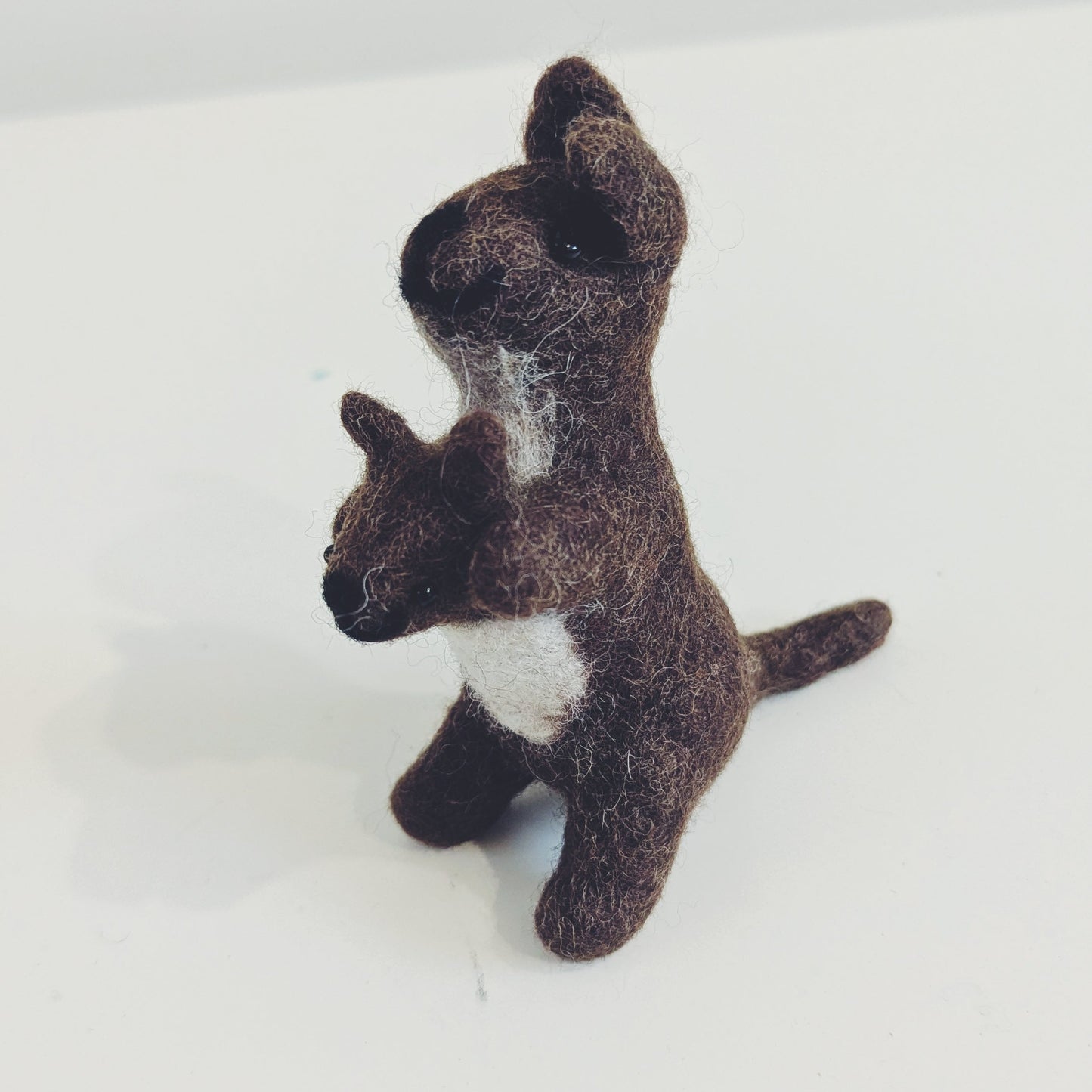 A small felted wool toy kangaroo holding a matching joey, crafted in dark brown with a white chest patch, standing on a plain white surface.