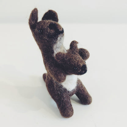 A small felted wool toy kangaroo holding a matching joey, crafted in dark brown with a white chest patch, standing on a plain white surface.