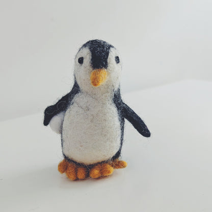 A small felt penguin figurine with black and white feathers, a yellow beak, and orange feet, standing on a plain white surface against a white background.