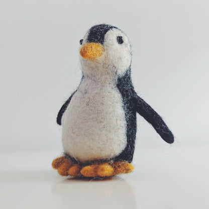A small felt penguin figurine with black and white feathers, a yellow beak, and orange feet, standing on a light gray surface against a pale gray background.