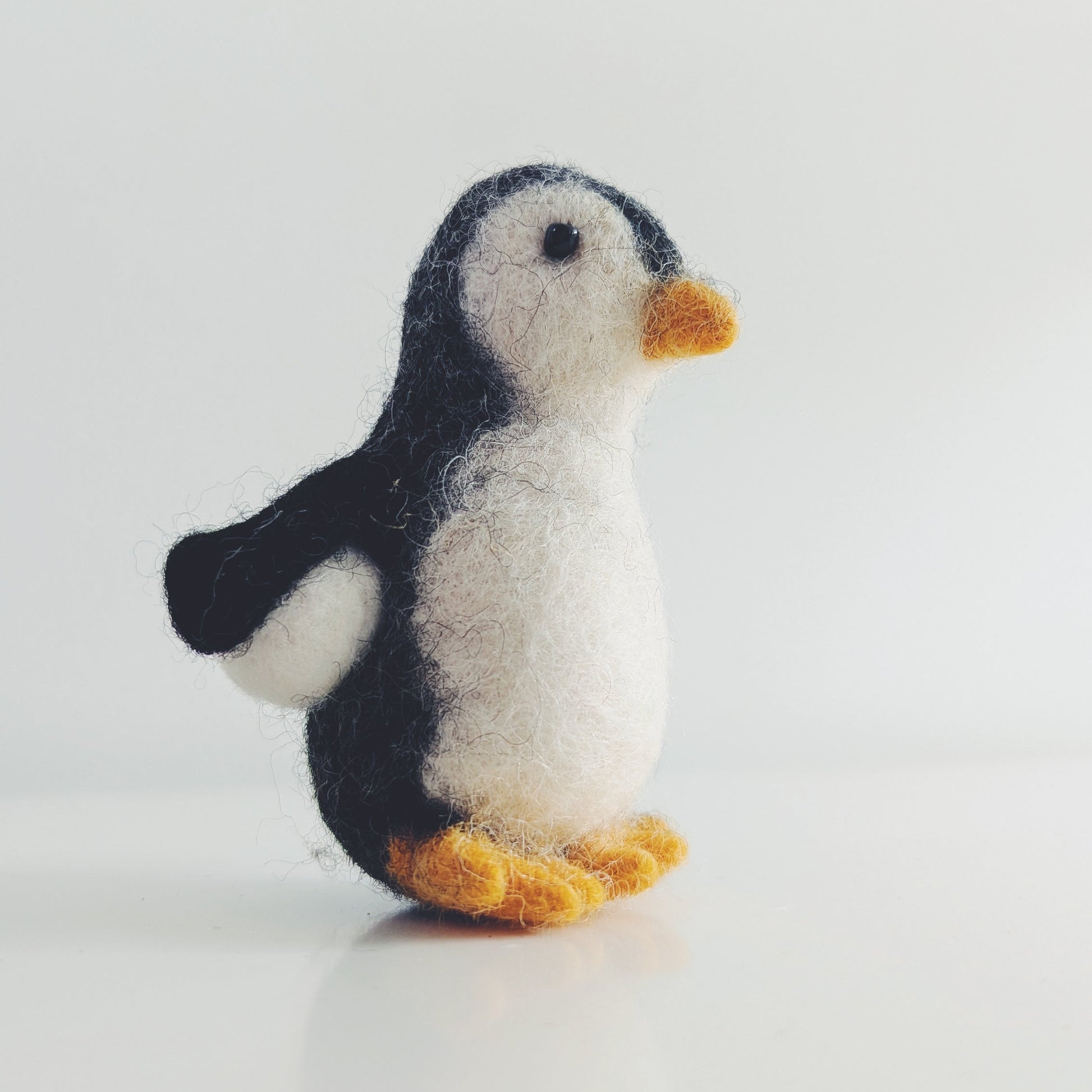 A small felt penguin toy with black and white feathers and orange beak and feet, standing on a light gray surface against a light gray background.