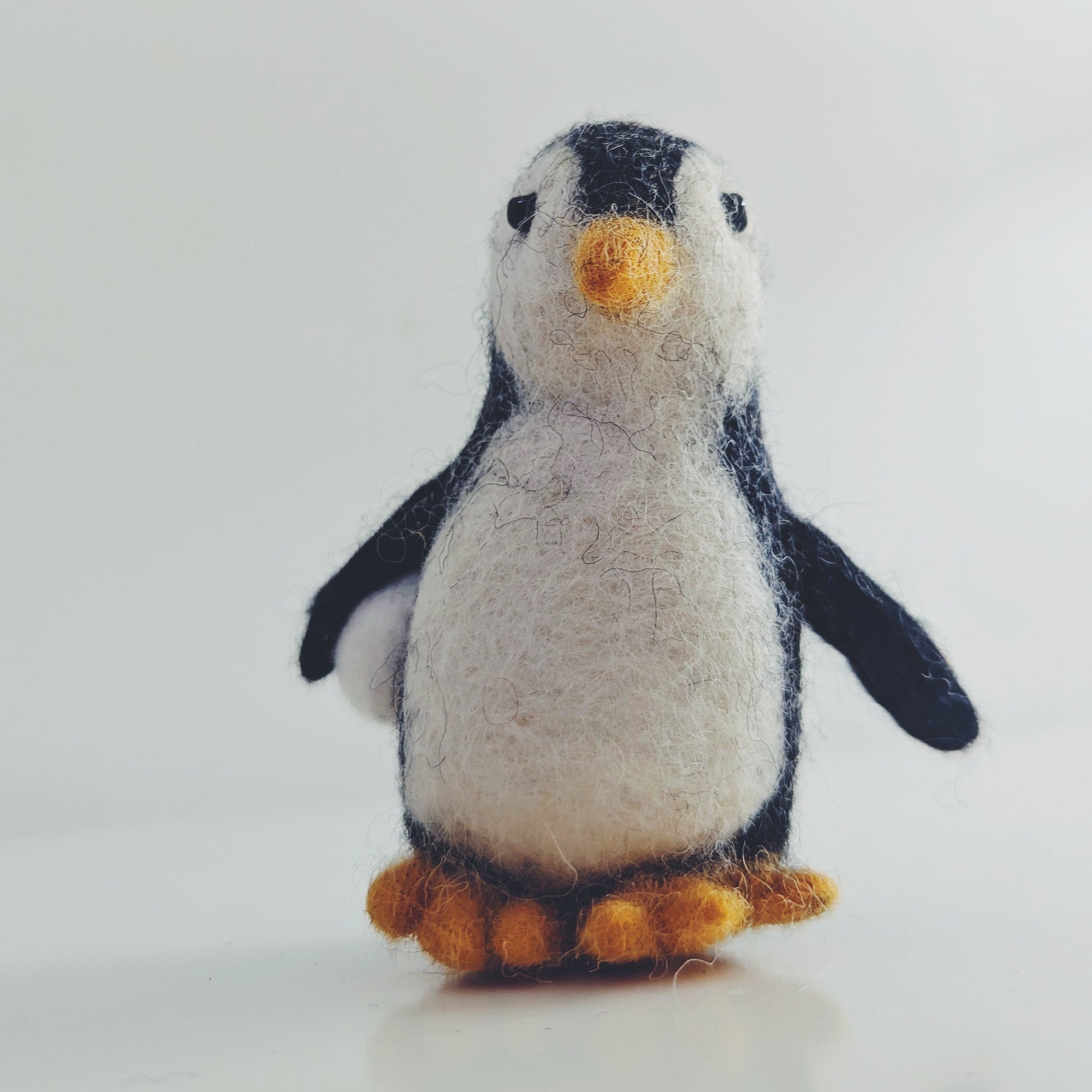 A small felt penguin toy standing upright on a light gray surface against a pale gray background. The penguin has a white body, black wings and head, a yellow beak, and orange feet.