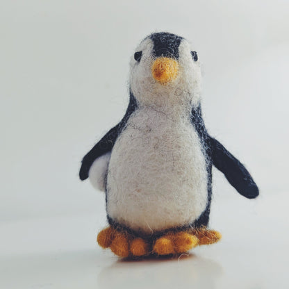 A small felt penguin toy standing upright on a light gray surface against a pale gray background. The penguin has a white body, black wings and head, a yellow beak, and orange feet.