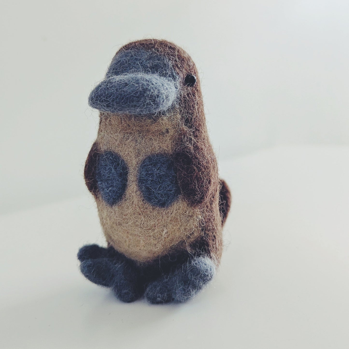 A small felted wool platypus figurine with a brown body, dark blue beak and feet, and dark blue spots on its back, standing on a white surface against a white background.