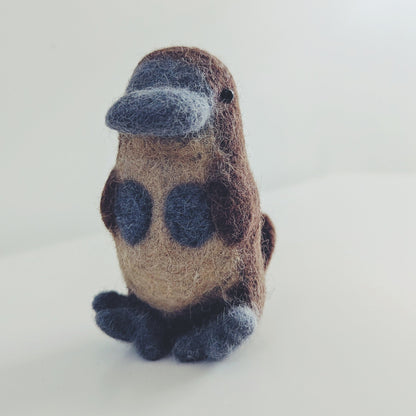A small felted wool platypus figurine with a brown body, dark blue beak and feet, and dark blue spots on its back, standing on a white surface against a white background.