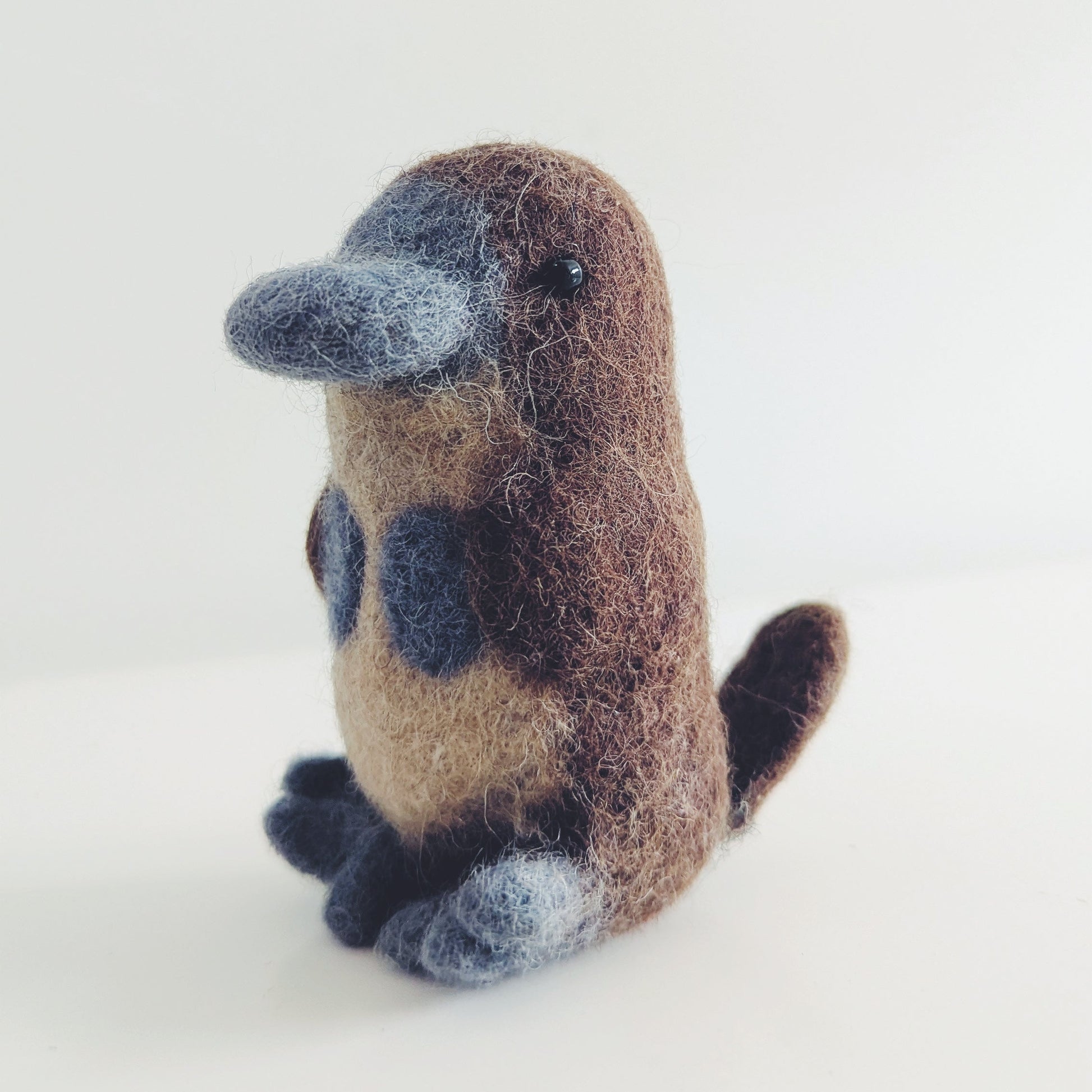 A small felted wool platypus figurine sitting upright on a plain white surface against a white background.