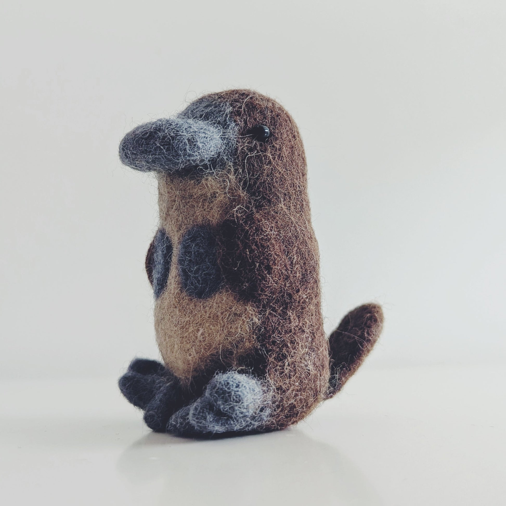 A small felted wool platypus toy figurine sitting upright on a clean white surface against a light gray background.