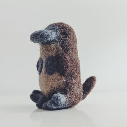 A small felted wool platypus toy figurine sitting upright on a clean white surface against a light gray background.