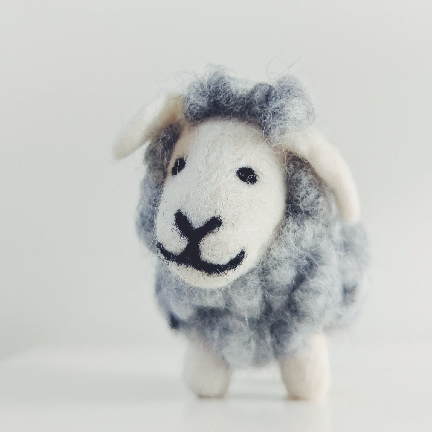 A small felted wool sheep figurine with a gray and white body, white face and ears, and black stitched eyes and mouth, standing on a white surface against a light gray background.