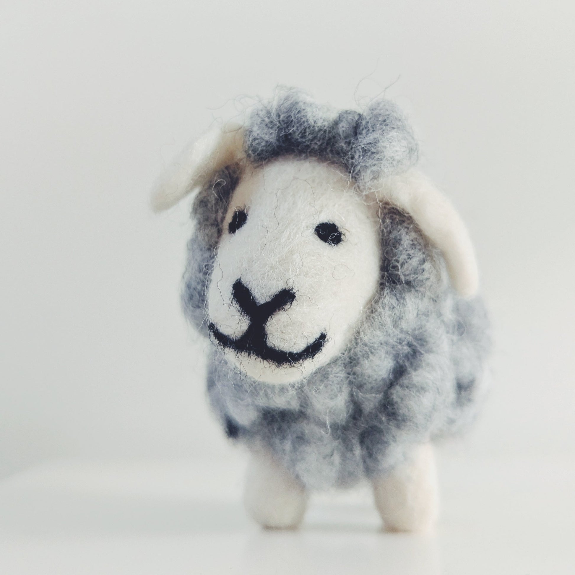 A small felted wool sheep figurine with a gray and white body, white face and ears, and black stitched eyes and mouth, standing on a white surface against a light gray background.