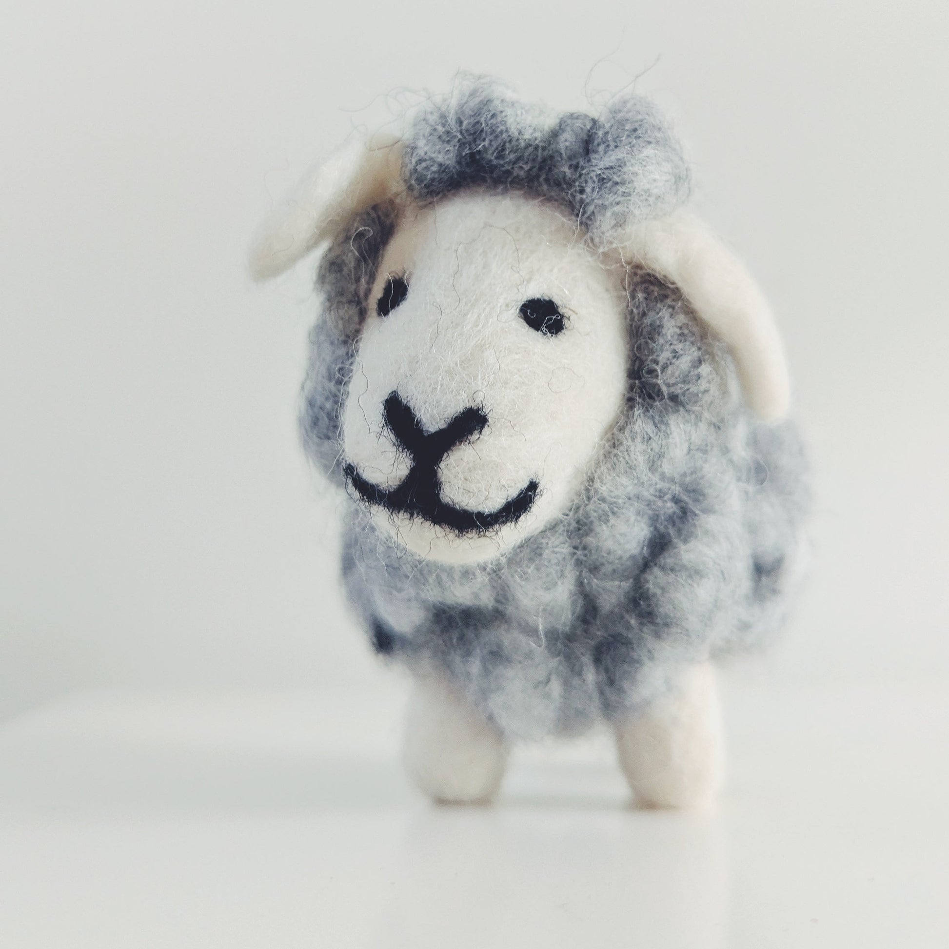 A small felted wool sheep figurine with a gray and white body, white face and ears, and black stitched eyes and mouth, standing on a white surface against a light gray background.
