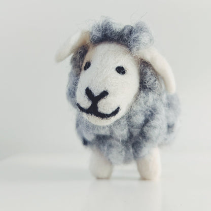 A small felted wool sheep figurine with a gray and white body, white face and ears, and black stitched eyes and mouth, standing on a white surface against a light gray background.