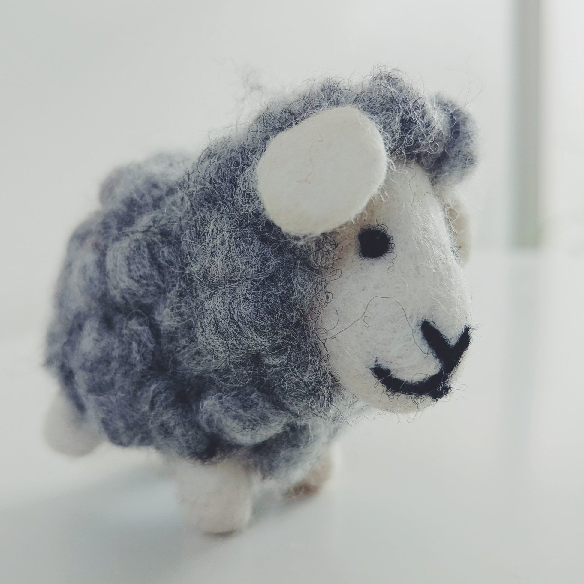 A small felted wool sheep figurine with a gray-blue woolly body, white face and ears, and black facial details, standing on a white surface against a soft white background.