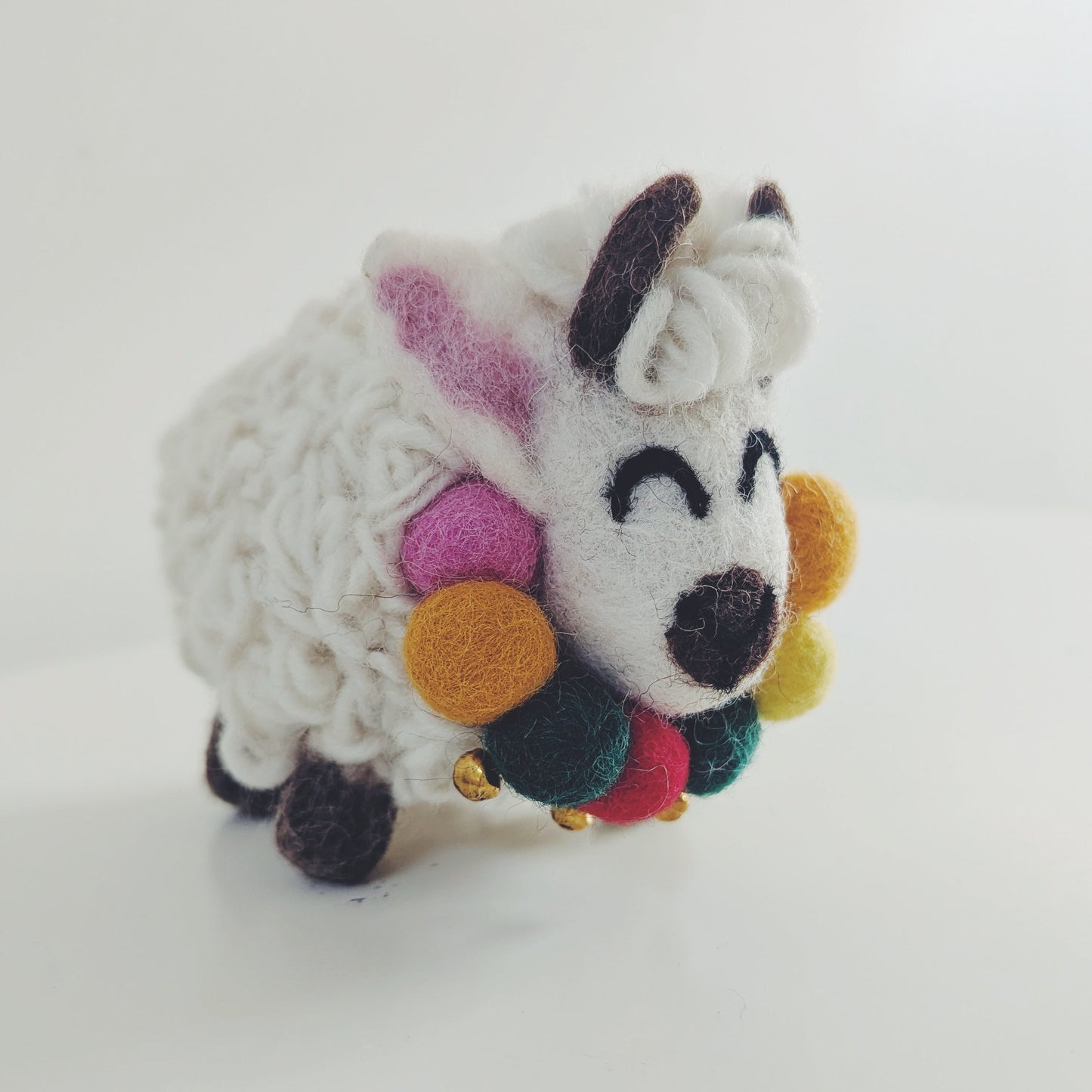 A small handmade felt yak figurine with a white textured body, black horns and hooves, pink inner ears, and colorful pom-pom accents on its face and neck, standing on a plain white surface.