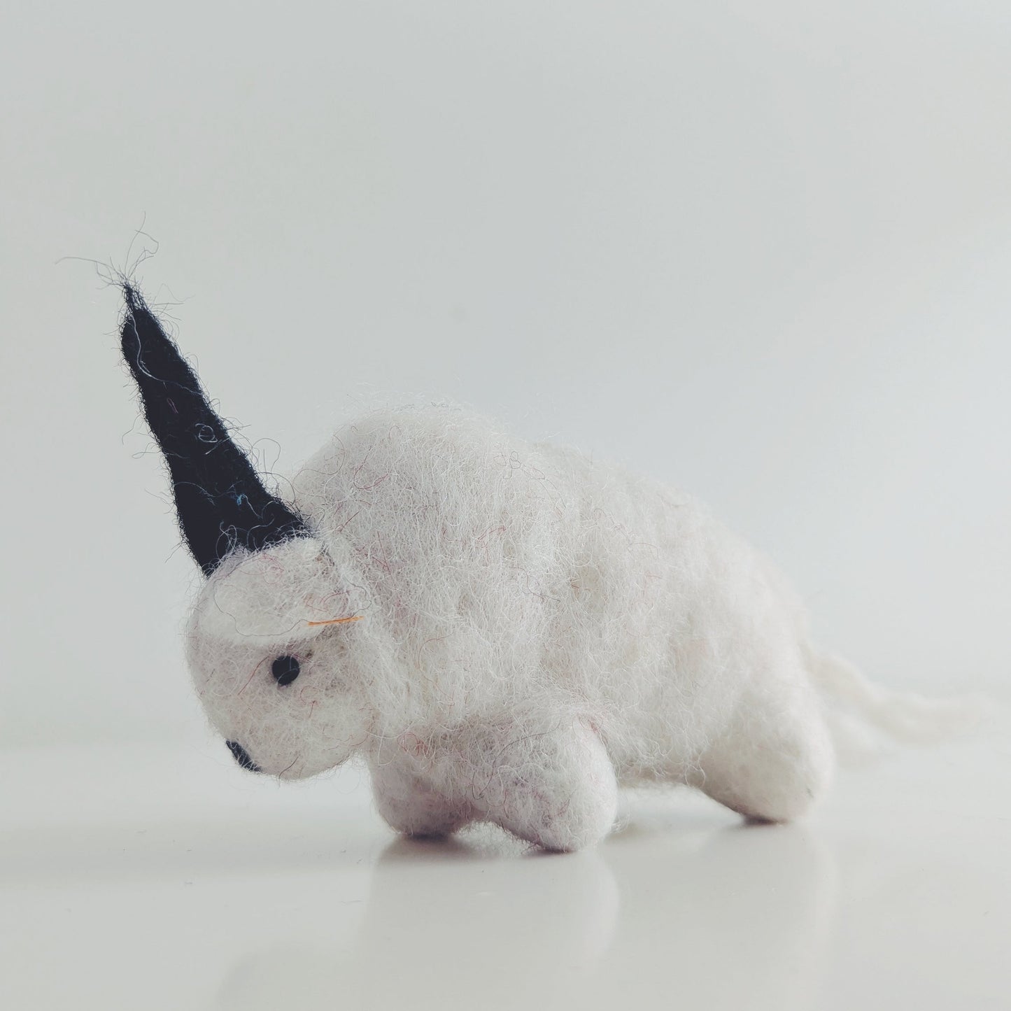 A small white felted himalayan goat figurine with a black horn or antler, photographed on a clean white studio background.