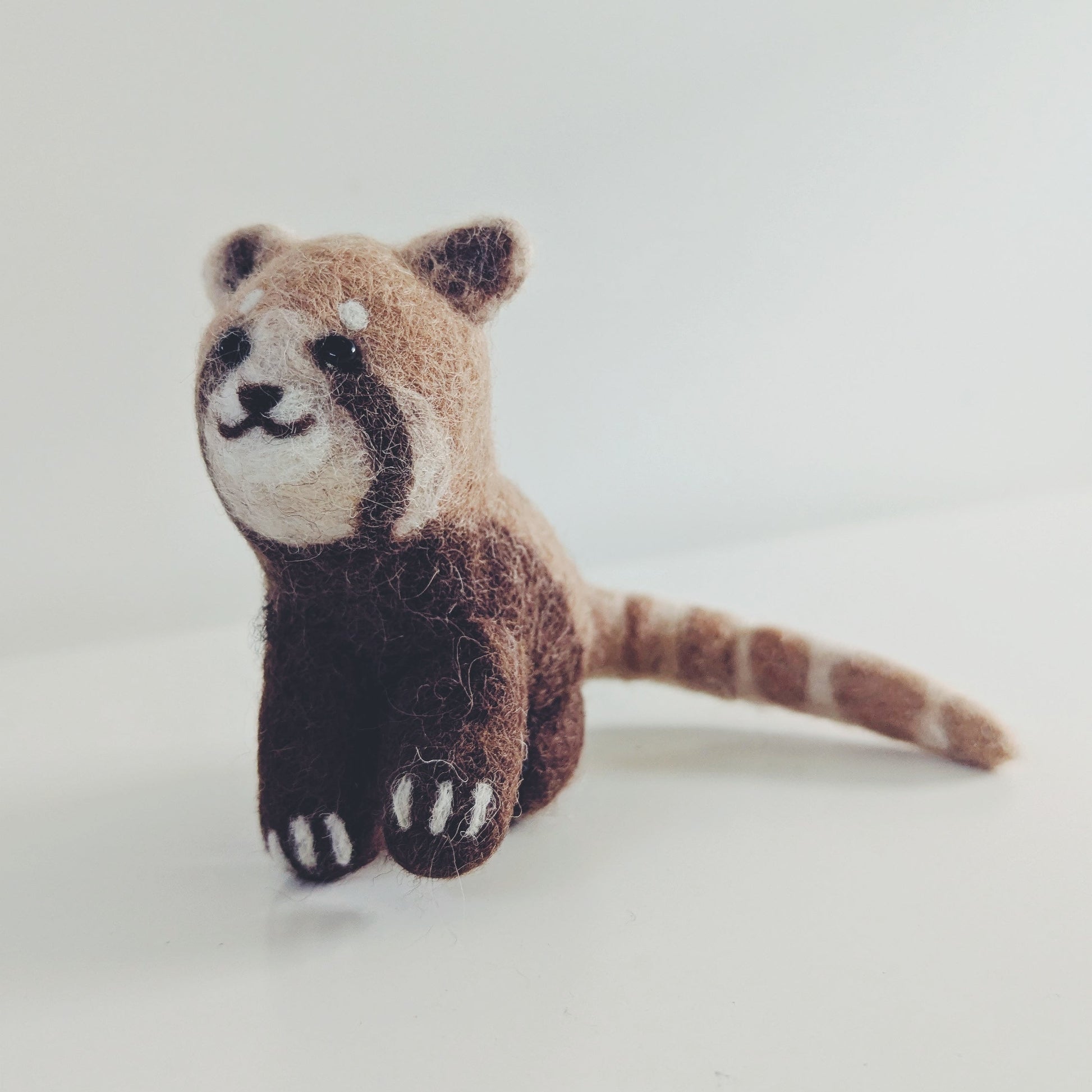 A small felted wool toy of a red panda sitting on a plain white background.