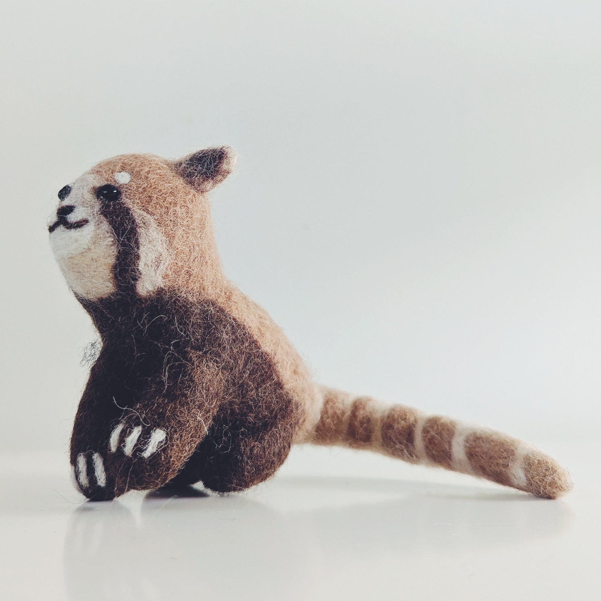 A small felted wool red panda toy sitting on a light gray reflective surface against a white background.