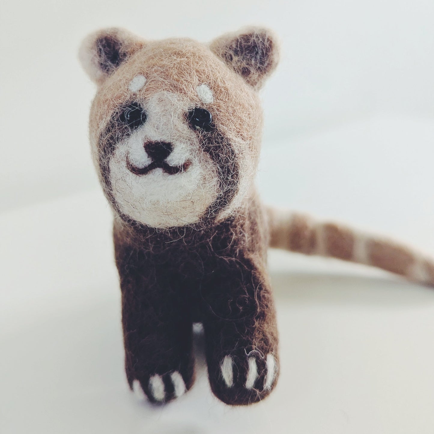 A small felted wool red panda figurine with brown, beige, and white colors, standing on a plain light background.