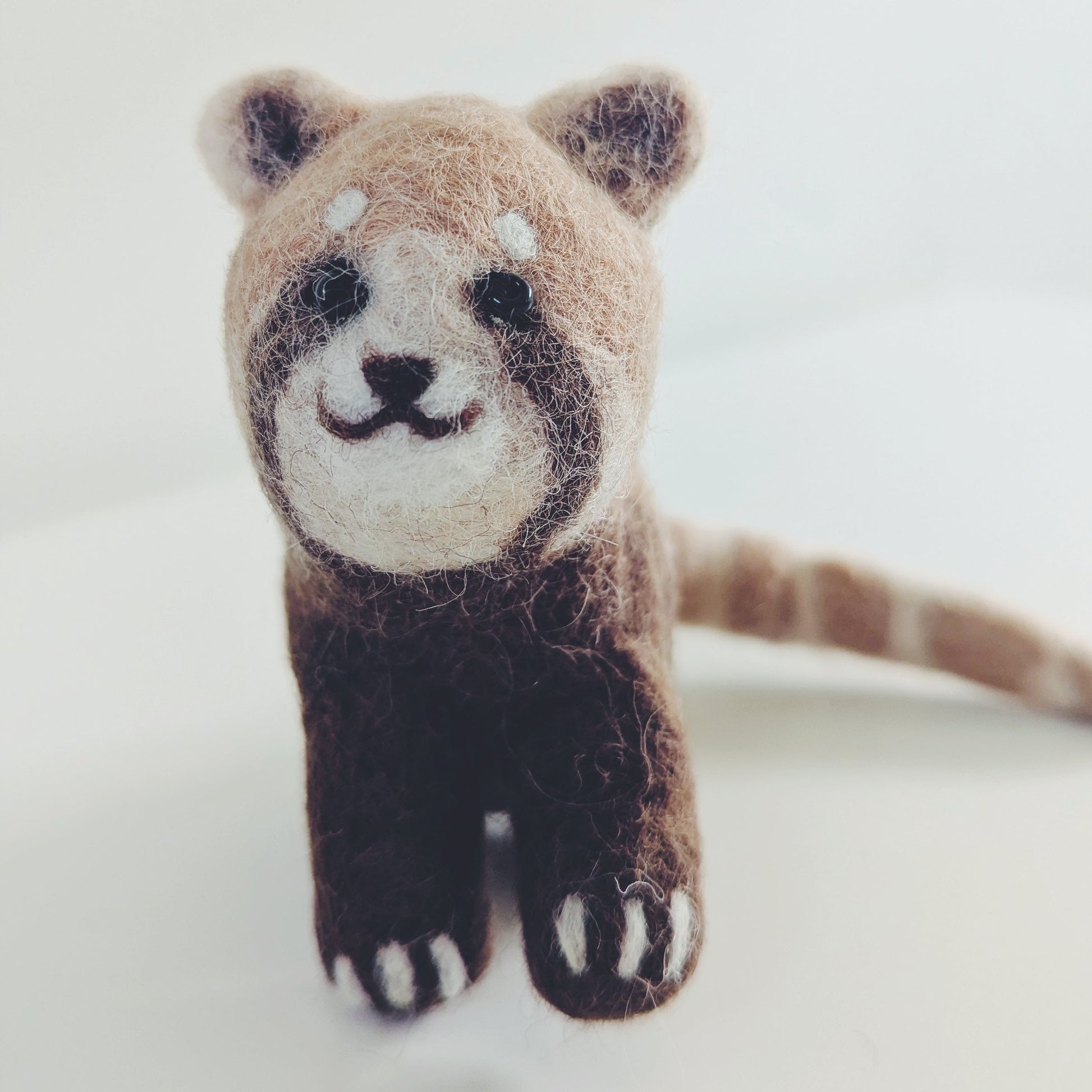 A small felted wool red panda figurine with brown, beige, and white colors, standing on a plain light background.
