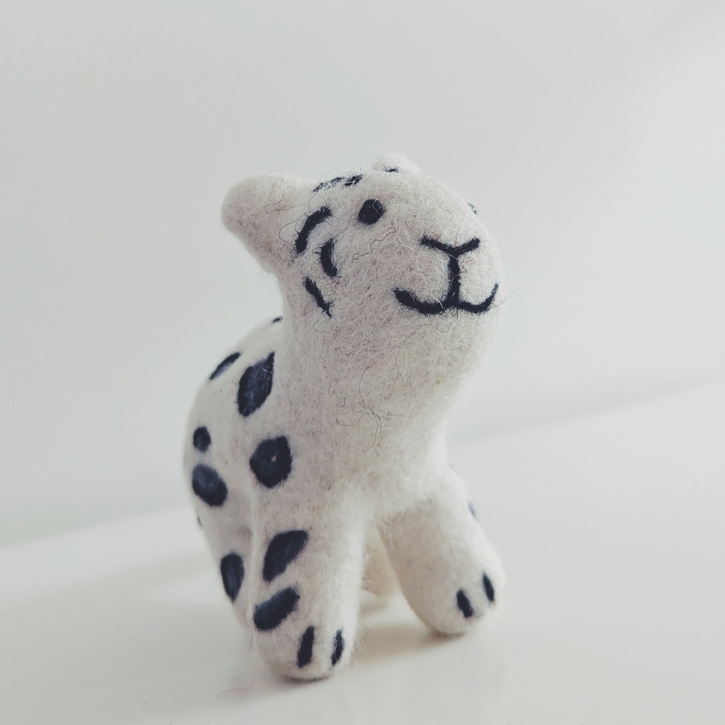 A small  felt snow leopard toy with black spots and a simple black stitched face, photographed on a plain light background.
