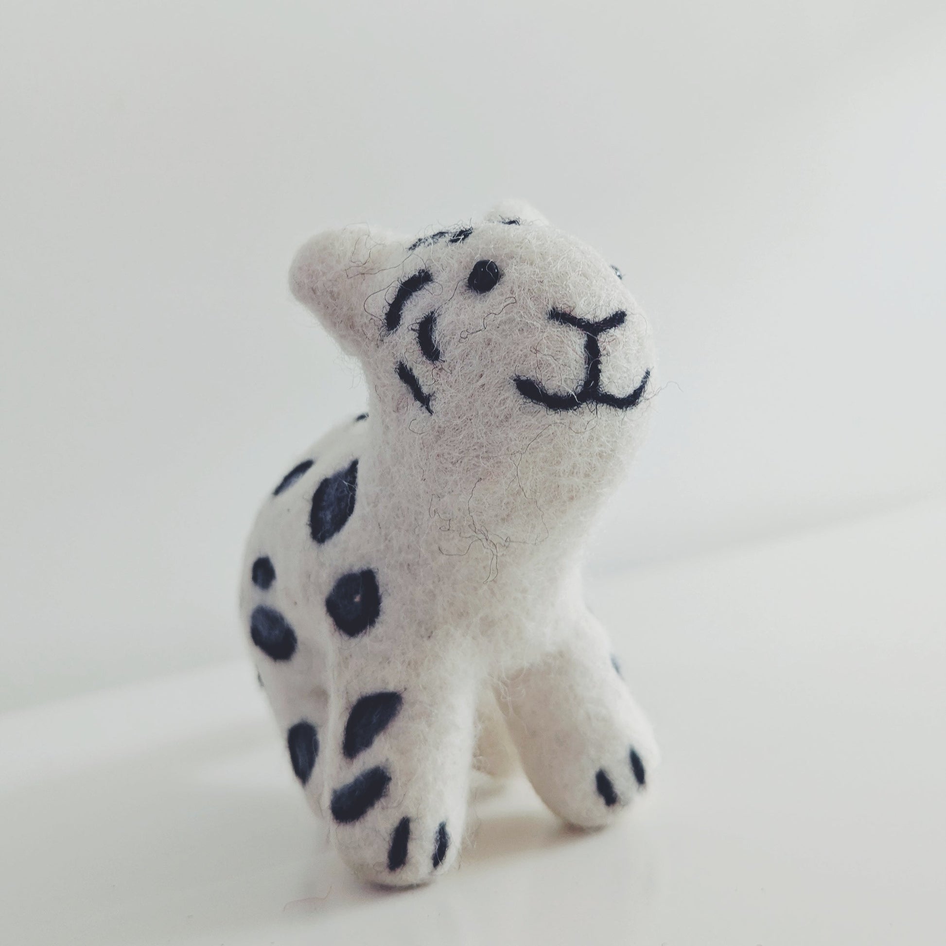 A small  felt snow leopard toy with black spots and a simple black stitched face, photographed on a plain light background.
