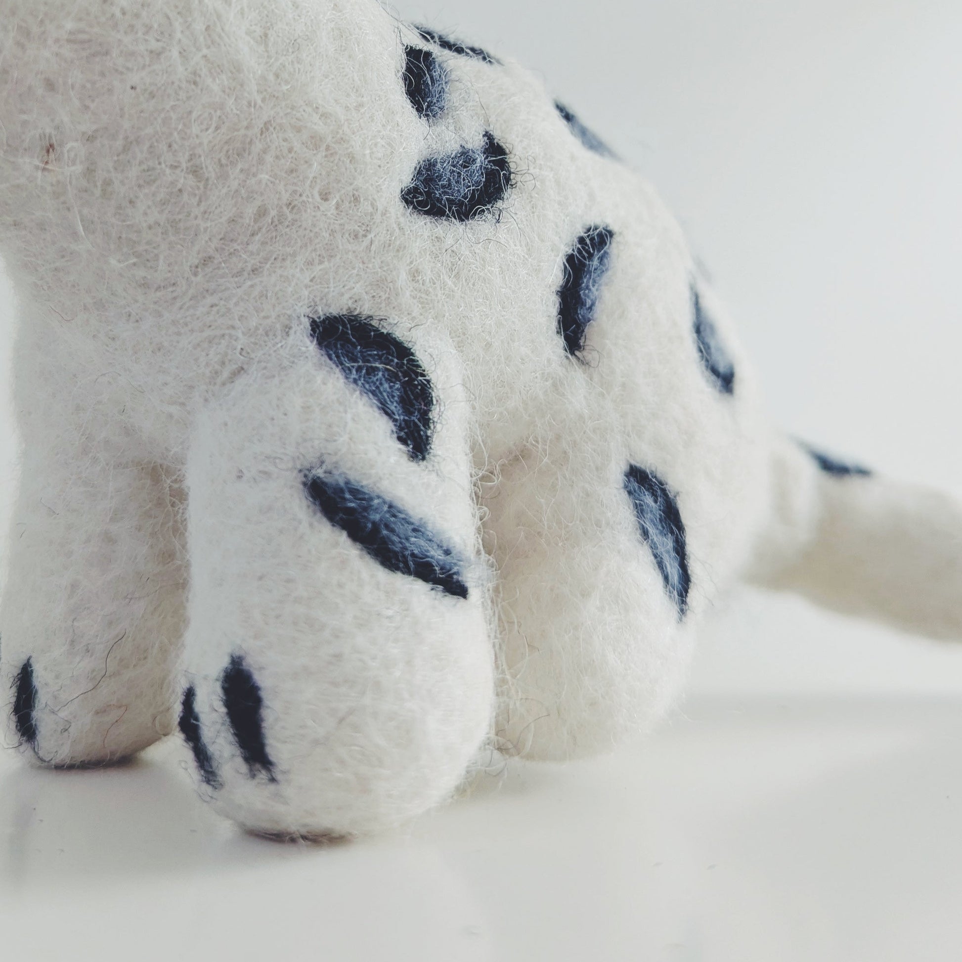 A white felted wool toy snow leopard cub with dark grey spots, photographed against a clean white background.