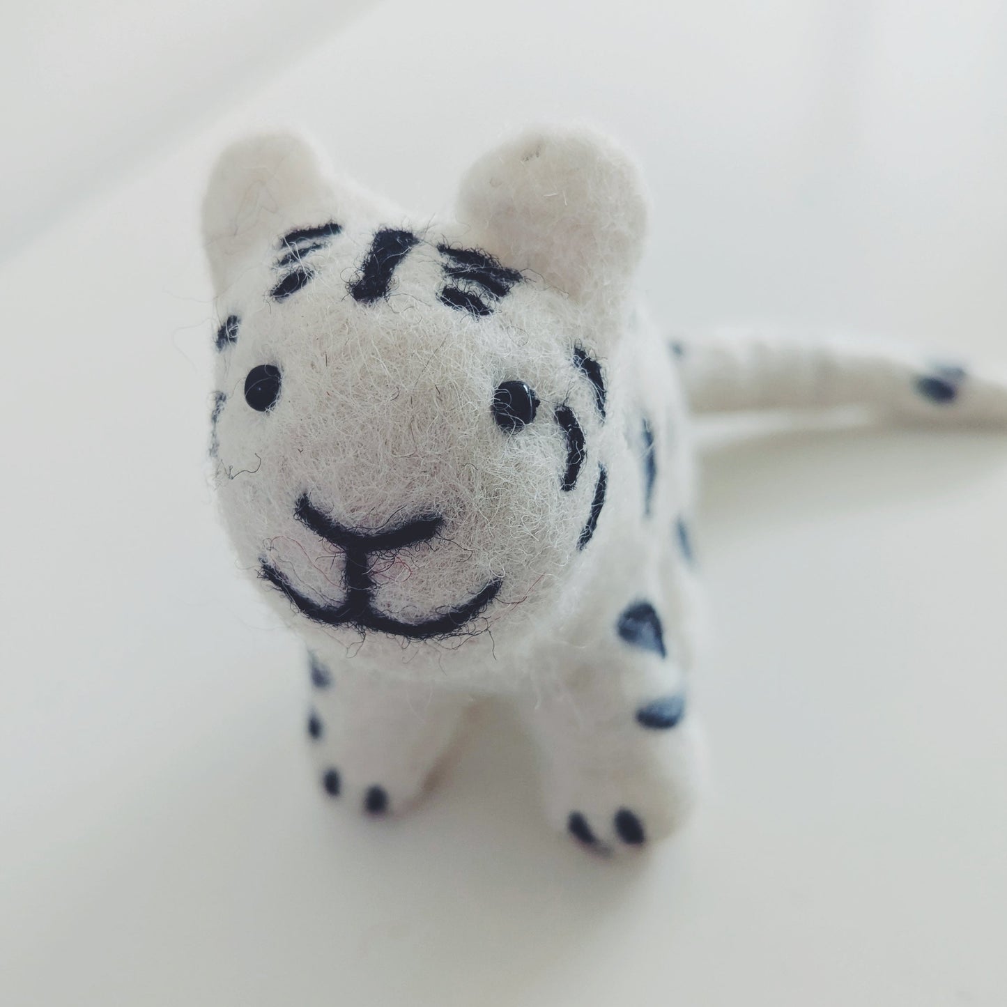 A small white felt snow leopard toy with black dry needled stripes and facial features, standing on a plain light surface.