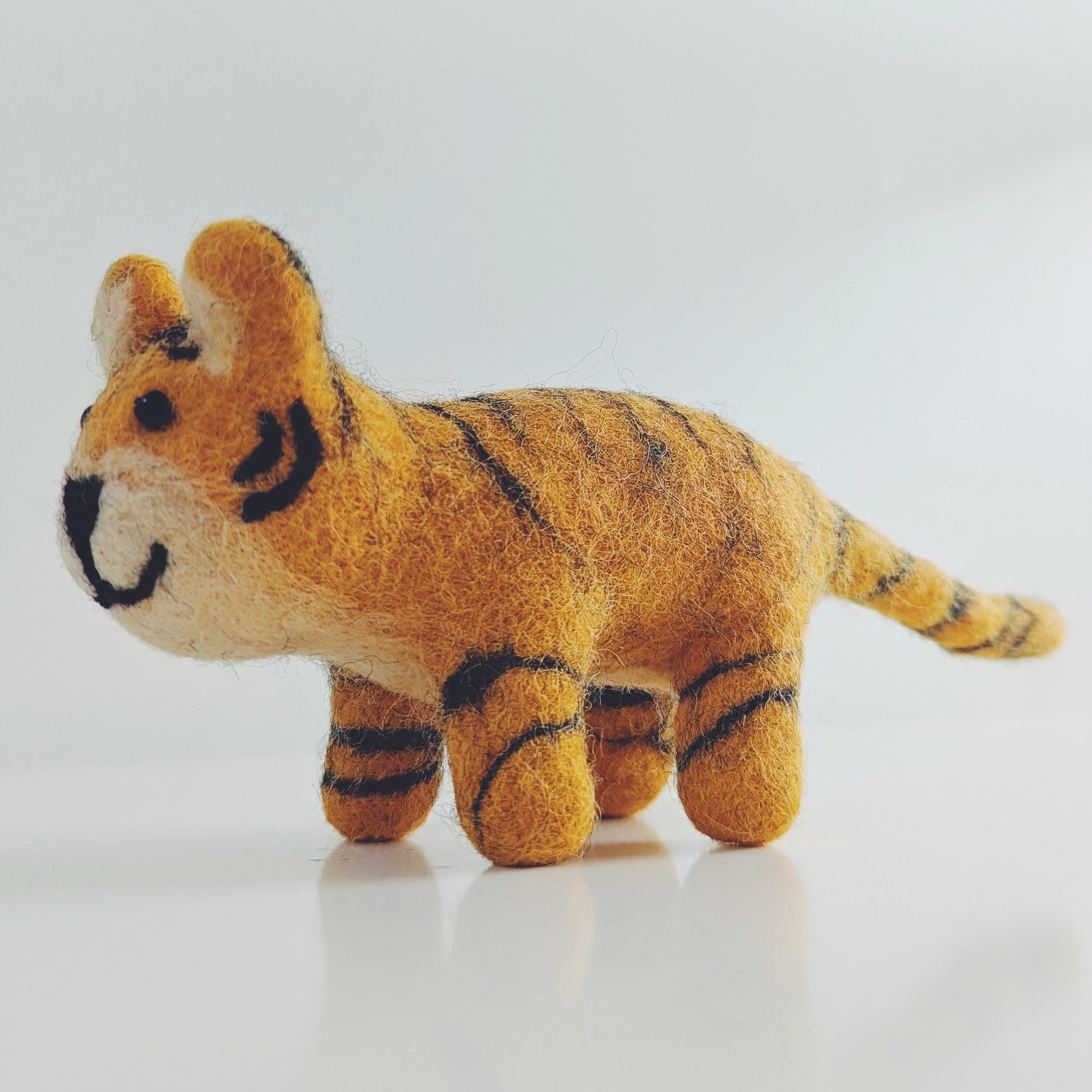 A small felt tiger toy with orange and black stripes, standing on a reflective white surface against a light gray background.