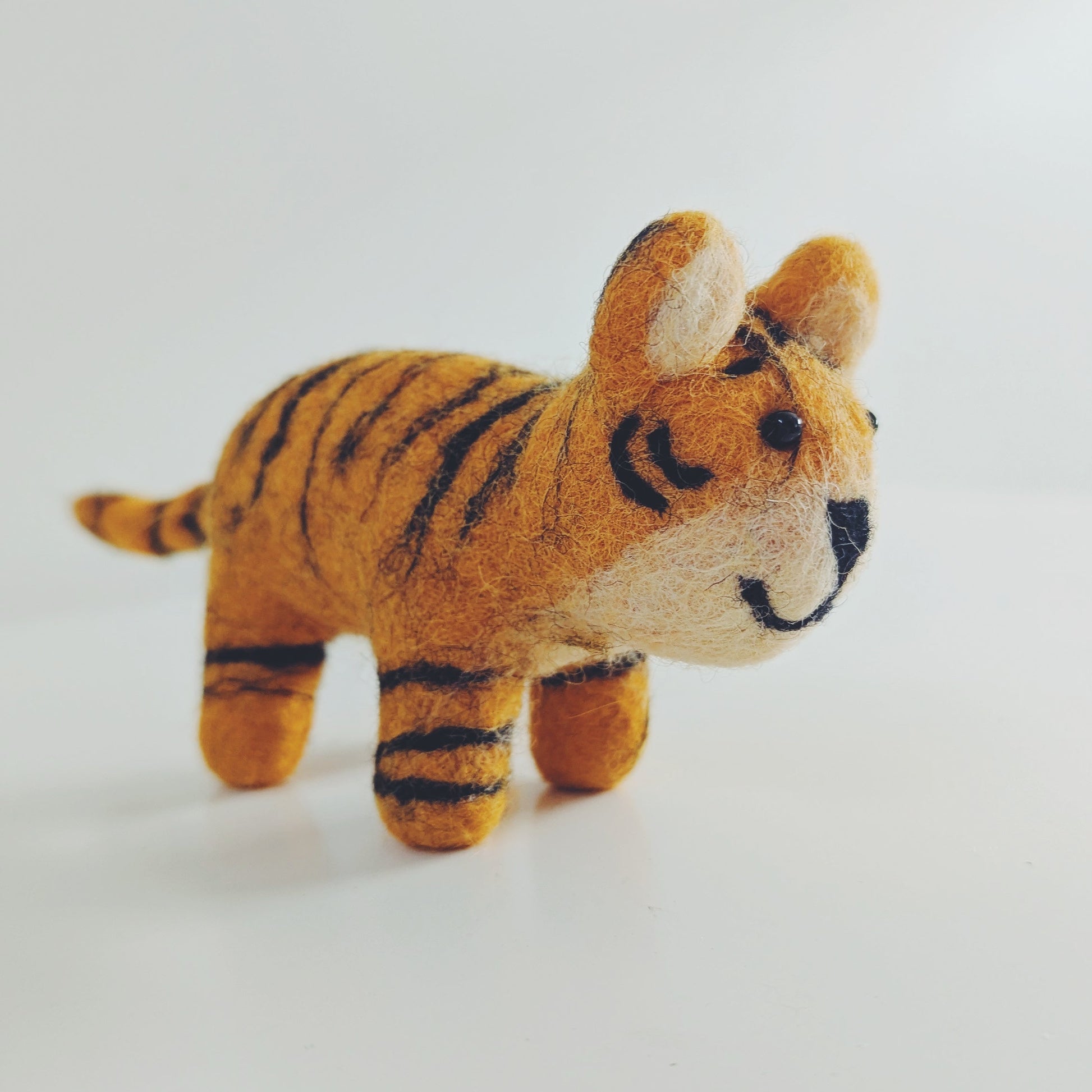 A small felt toy tiger figurine in orange and black stripes with a cream-colored face and paws, standing on a plain white surface against a white background.