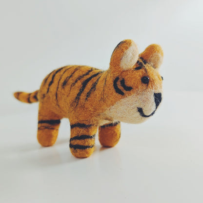 A small felt toy tiger figurine in orange and black stripes with a cream-colored face and paws, standing on a plain white surface against a white background.