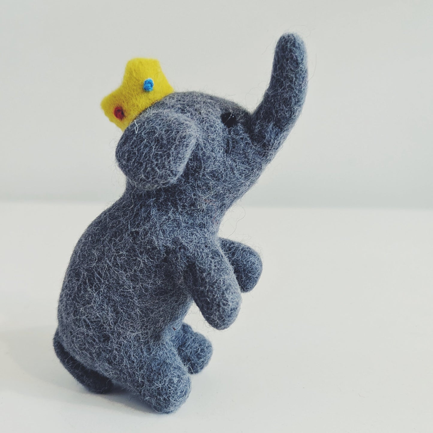 A small felt elephant figurine in dark blue-gray, wearing a tiny yellow crown with red and blue details, photographed on a plain light background.