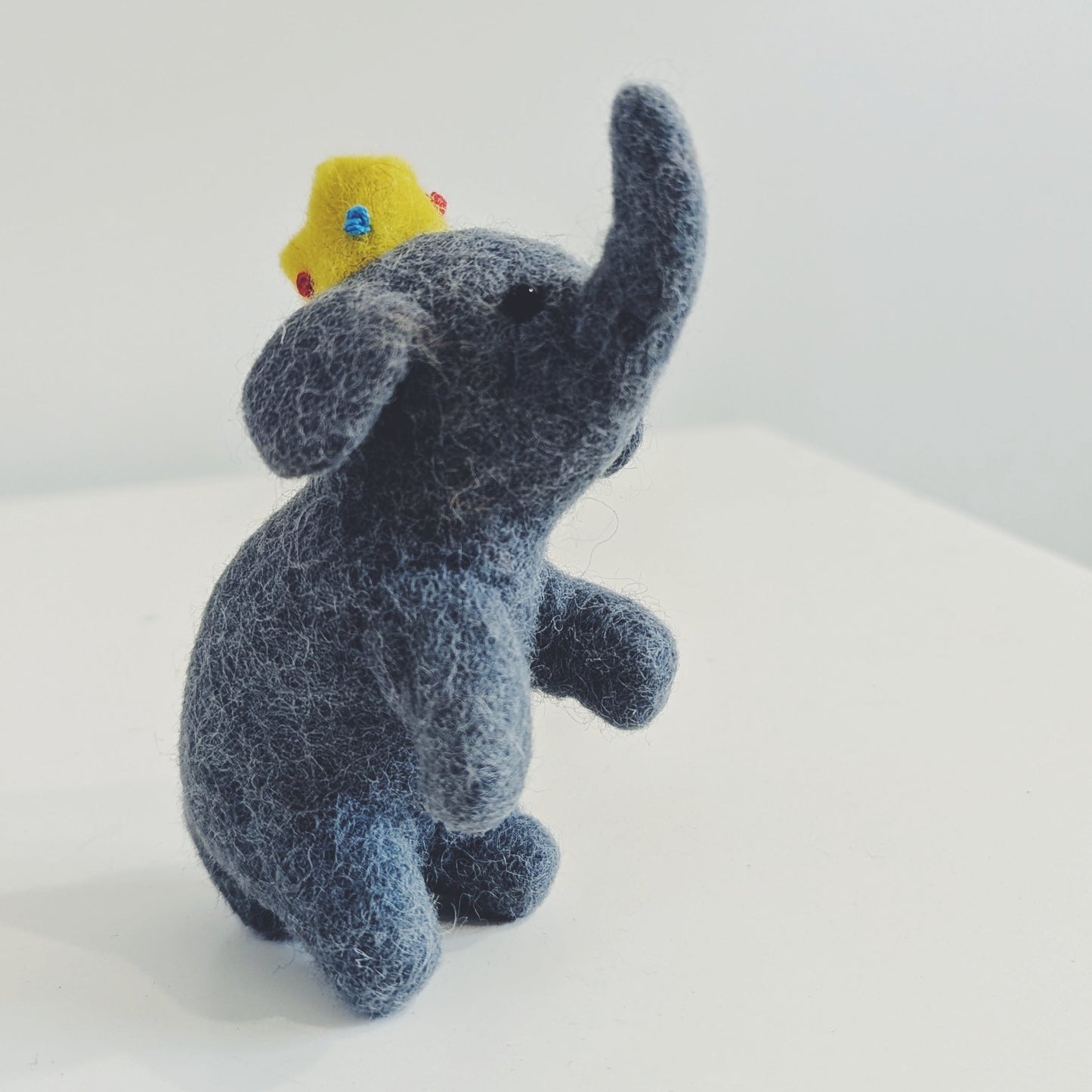 A small felt elephant toy in dark blue-gray with a small yellow duck figure on its back, photographed on a plain white surface against a white background.