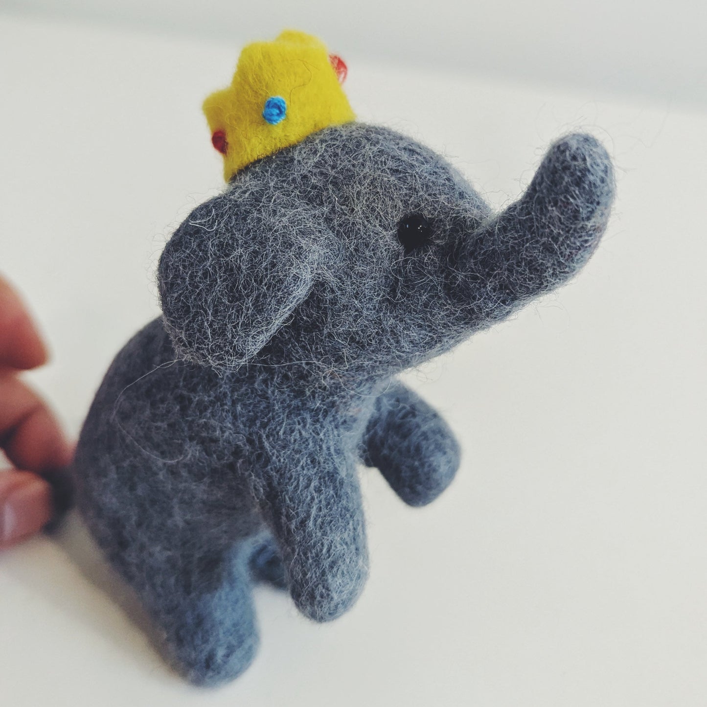 A small felt elephant figurine in dark blue-gray with a yellow crown, photographed on a plain light background. A human hand is visible at the left edge holding the item.