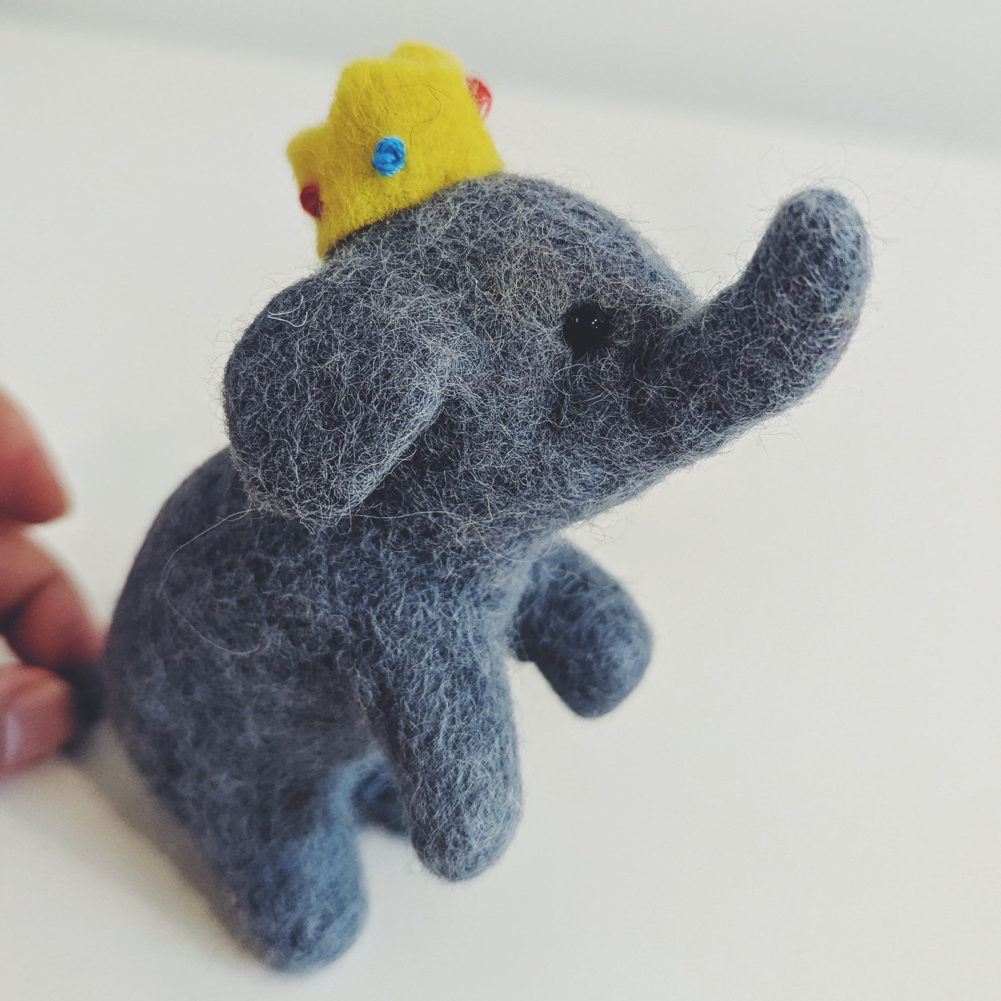 A small felt elephant figurine in dark blue-gray with a yellow crown, photographed on a plain light background. A human hand is visible at the left edge holding the item.