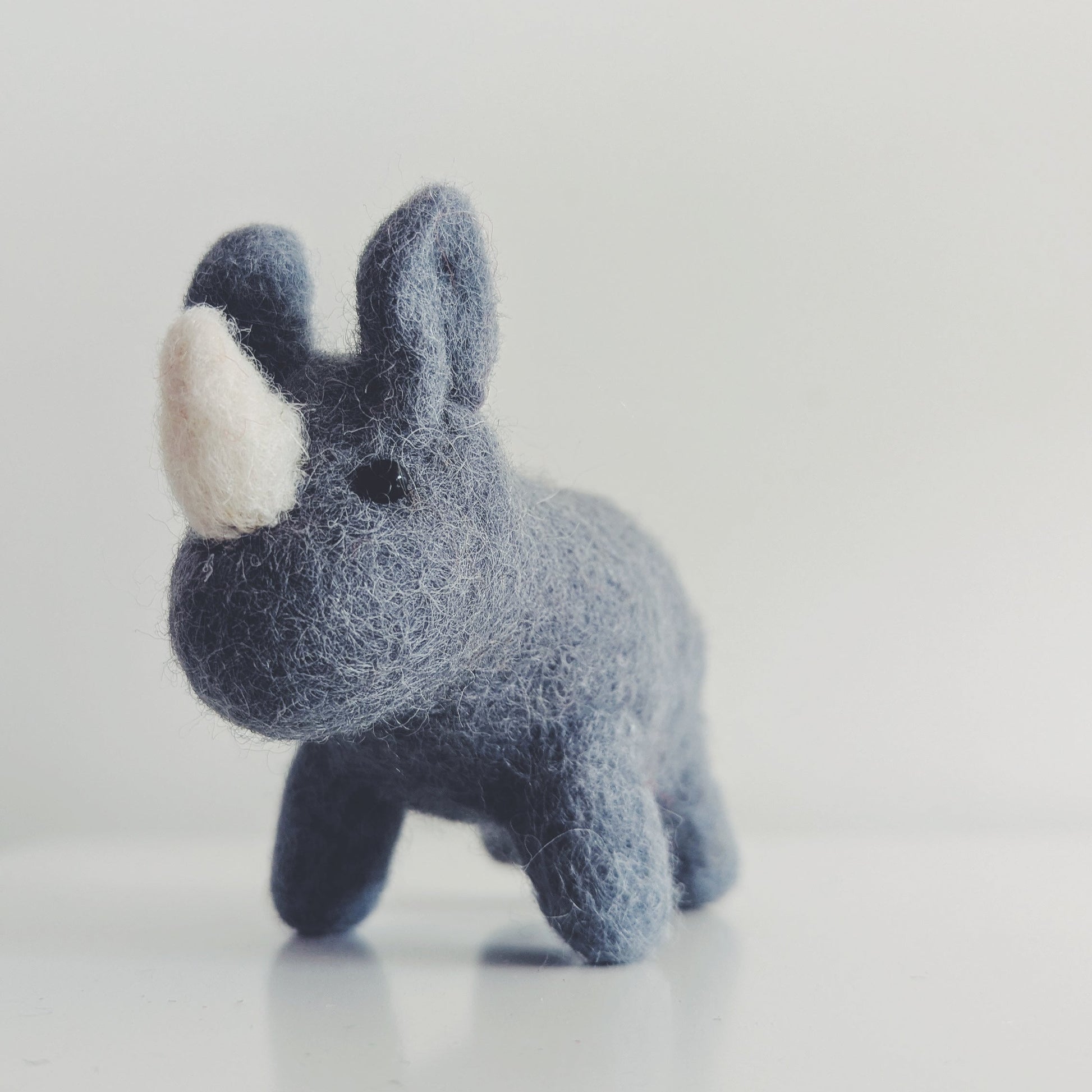 A small felt rhinoceros toy in dark blue-gray with a white horn, standing on a light gray reflective surface.